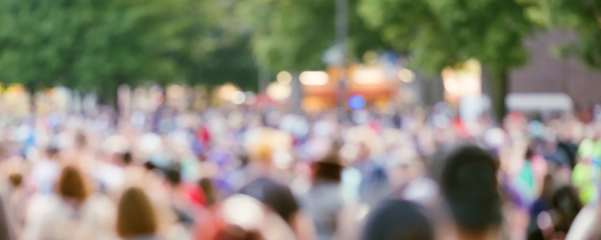 A crowd filling a tree-lined street at an outdoor event, shot out of focus so no one person is identifiable.
