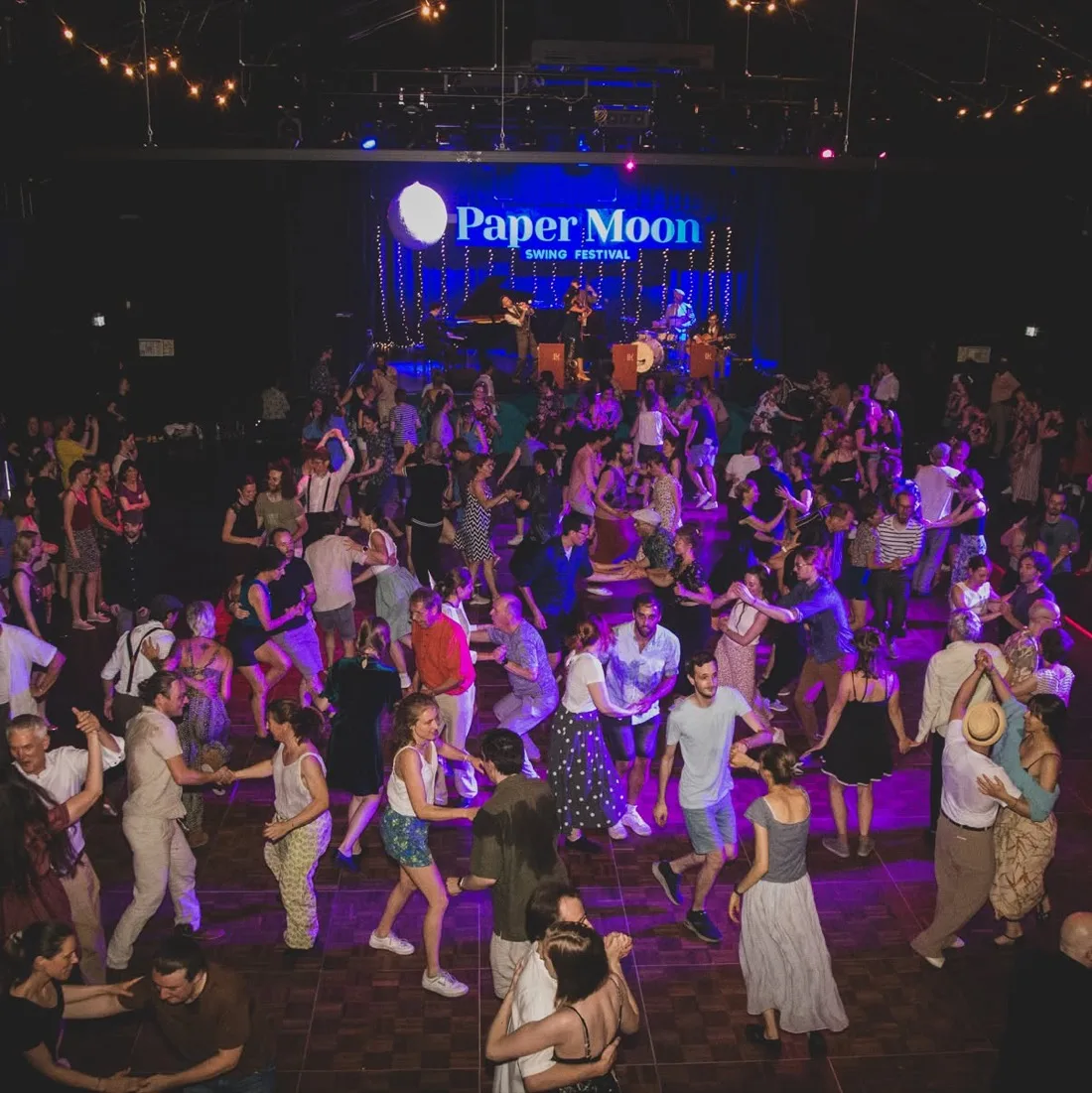 The social dance floor at Paper Moon: a crowded hall of couples dancing under string lights, with the band playing on a blue-lit stage beneath the festival's paper-moon backdrop.